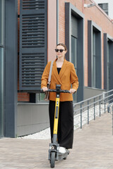 Poster - Young female broker in sunglasses and formalwear standing on electric scooter and riding home after work while moving along street