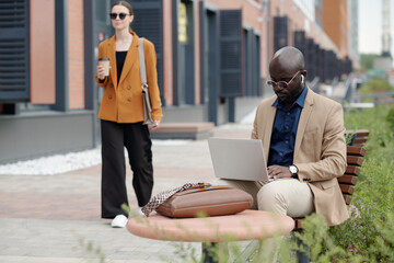Aufkleber - Serious African American businessman in earphones typing on laptop keyboard while sitting on bench against young woman with cup of coffee