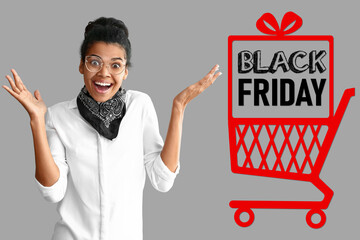 Surprised African-American woman, drawn shopping cart and text BLACK FRIDAY on grey background