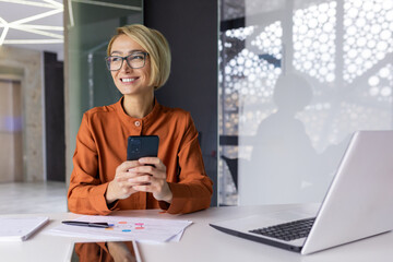 Young beautiful woman at workplace using application on phone, business woman dreamy thinking looking out window holding smartphone, working inside office with laptop.