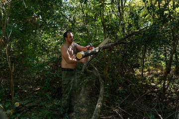 A young lumberjack cuts a fallen branch in the forest on a hot summer day. He uses a chainsaw, wearing camo pants and no shirt due to the heat and humidity