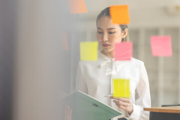 business asian girl in white shirt thinks, holds pen and clipboard, writing sticking adhesive notes on glass wall, making notes, standing over white background