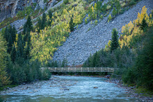 Scenic View Of A Bridge Crossing A Flowing River While Yellow Aspen Trees Glow On The Hillside Among Falling Rock