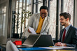 © La Famiglia - A female executive and her male colleague, representing different ethnicities, engage in a focused discussion at their office workstation, leveraging technology for effective teamwork
