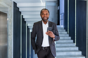Portrait of happy successful African American boss, man smiling and looking at camera, businessman holding phone using online application, man walking down corridor of building inside office.