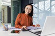 © Liubomir - Portrait of young Asian successful financier working on paperwork with calculator inside office, businesswoman smiling and looking at camera, filling forms and verifying financial reports.