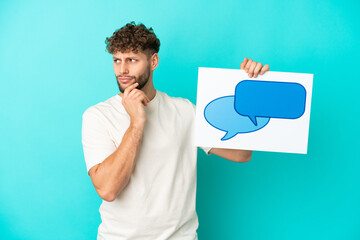 Wall Mural - Young handsome caucasian man isolated on blue background holding a placard with speech bubble icon and thinking