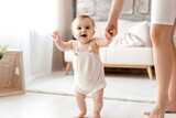 Fototapeta  - charming little girl in diapers takes her first steps at home holding hands with her mother, the child learns to walk at home in a bright living room