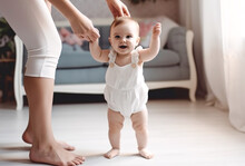 Charming Little Girl In Diapers Takes Her First Steps At Home Holding Hands With Her Mother, The Child Learns To Walk At Home In A Bright Living Room