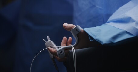 Male patient lies on table under anesthesia in surgery room. Surgeons perform difficult operation. Close up of hand of man with medical sensors and pulse meter. Modern equipment in clinic or hospital.