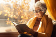 Beautiful senior lady reading a book by a window on sunny autumn day. Elderly woman enjoying nice fall weather.