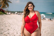 Happy sexy curvy plus size model in a red one-piece swimsuit on the beach, fat busty chubby woman resting at the sea on summer holidays