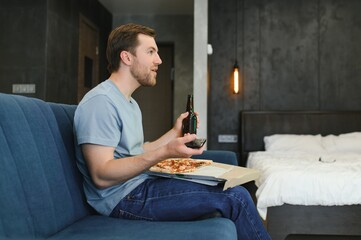 Happy young man drinking beer and eating pizza when watching game on tv at home