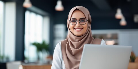 Portrait of a smiley young Muslim woman in eyeglasses wearing hijab working with laptop computer in her modern business office.