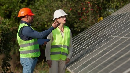 Wall Mural - A man and a woman in hard hats and with a laptop are discussing work tasks against the backdrop of solar panels outside. Female environmental engineer talking to investor. Green electricity concept.