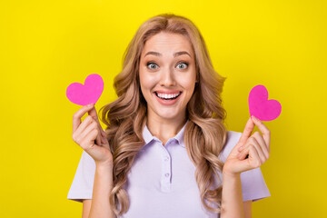 Sticker - Photo of adorable impressed girl with curly hairstyle wear violet polo finger hold two small hearts isolated on yellow color background