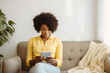 © Jasmina - Beautiful smiling black woman siting on the couch and scrolling on her phone at home