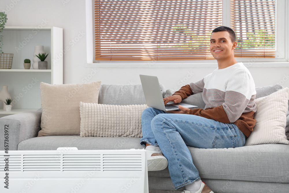 Young man with laptop warming near radiator at home