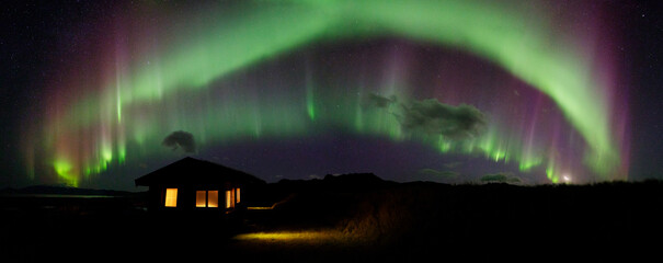 Wall Mural - a panorama of aurora borealis over a cabin with glowing lights illuminating the tall grass on a clea