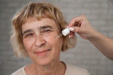 Wall Mural - Close-up portrait of an old woman applying hyaluronic acid serum with a pipette. Anti-aging face care. 