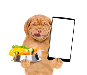 Wall Mural - Happy Licking lips puppy holding bowl of vegetables and shows empty screen of smartphone. isolated on white background