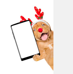 Wall Mural - Happy Mastiff puppy dressed like santa claus reindeer  Rudolf holds big smartphone with empty screen in it paw, showing close to camera behind empty white banner. isolated on white background