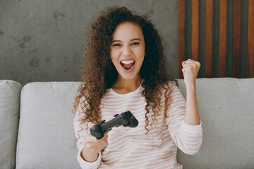 Young woman wear casual clothes sit on grey sofa couch hold in hand play pc game with joystick console do winner gesture stay at home hotel flat rest relax spend free spare time in living room indoor