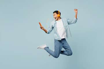 Wall Mural - Full body young man of African American ethnicity he wear shirt casual clothes jump high listen to music in headphones use mobile cell phone isolated on plain pastel light blue cyan background studio.