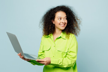 Wall Mural - Side view smiling young IT latin woman she wear green shirt casual clothes hold use work on laptop pc computer look aside isolated on plain pastel light blue cyan background studio. Lifestyle concept.