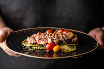 A man in black holds a roasted duck breast fillet on caramelized apples on a black background