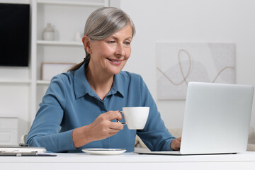 Poster - Beautiful senior woman with cup of drink using laptop at white table indoors