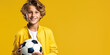 Boy holding a ball and smiling against a yellow studio background, wearing a long yellow jacket and a white inner shirt. Space for copy paste