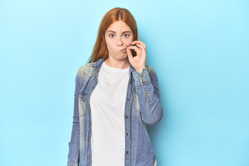 Wall Mural - Redhead young woman on blue background with fingers on lips keeping a secret.