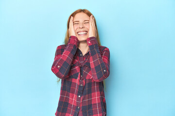 Wall Mural - Redhead young woman on blue background laughs joyfully keeping hands on head. Happiness concept.