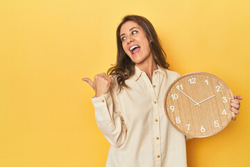 Wall Mural - Woman holding clock on yellow backdrop points with thumb finger away, laughing and carefree.