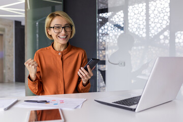 Portrait of successful woman winner, businesswoman smiling and looking at camera, worker holding phone in hands received notification of successful contract approval, celebrating holding hand up.