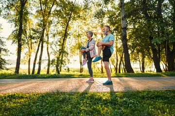Wall Mural - Healthy seniors working out outside. Side view of two dynamic family partners standing on pathway in park and doing stretching exercise for legs. Man and woman lifting and clutching knees to abdomen.