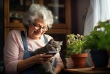 Fototapeta  - An elderly woman at home sitting on the sofa with her pet cat is holding a smartphone and perhaps taking a selfie with her furry friend.