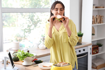 Wall Mural - Beautiful young woman eating tasty sandwich in kitchen