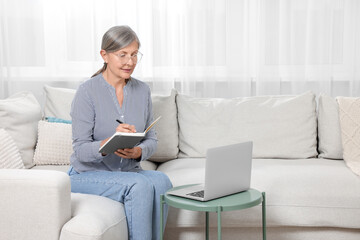 Wall Mural - Beautiful senior woman writing something in notebook while working with laptop at home