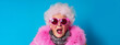 Colorful studio portrait of eccentric elderly granny wearing pink fur and sunglasses, blue background, shocked and judgmental expression