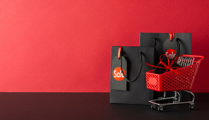 Wall Mural - Black Friday haul: Side view of tiny shopping cart with gift box and black paper bags, price tags displayed on a black table. Red wall backdrop, perfect for text or ads