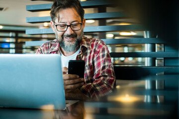 Happy modern mature man at work in coworking workplace alternative office using laptop computer and mobile phone to communicate. Devices connection. Adult male smile and use smartphone app indoor