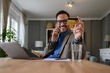 Wall Mural - Handsome entrepreneur in elegant suit holding credit card and talking over smart phone at desk