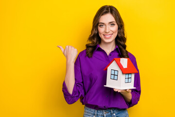 Wall Mural - Portrait of charming cheerful person hold small house indicate finger empty space isolated on yellow color background