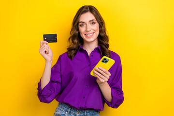 Poster - Photo of toothy beaming positive woman with wavy hair dressed purple shirt hold debit card smartphone isolated on yellow background