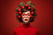Funny woman portrait on red background, fun hair with fashion hat made of Christmas tree branches, balls and baubles, red glasses and lips, standing out of the crowd, different for holidays concept