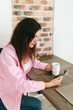© Mila - A young girl with black hair in a pink shirt sits at a table with a phone and a white mug in a cozy room with brown brick. Woman working, talking on the phone, drinking coffee in a modern cafe.Morning