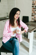 © Mila - A young girl with black hair in a pink shirt sits at a table with a phone and a white mug in a cozy room with brown brick. Woman working, talking on the phone, drinking coffee in a modern cafe.Morning