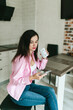 © Mila - A young girl with black hair in a pink shirt sits at a table with a phone and a white mug in a cozy room with brown brick. Woman working, talking on the phone, drinking coffee in a modern cafe.Morning
